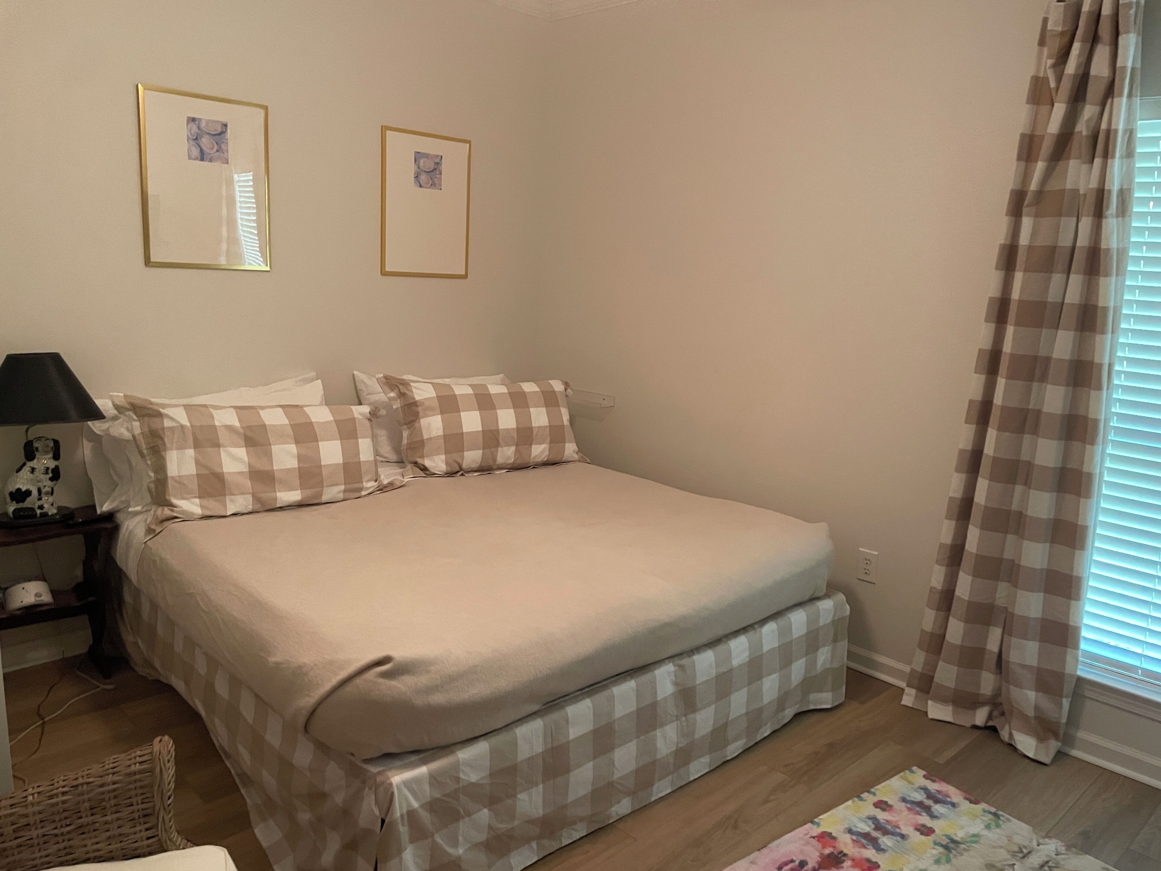 A bedroom with a bed, two framed pictures on the wall, a lamp, and a checkered bed skirt.