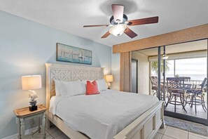 A hotel room with a bed, two bedside lamps, a ceiling fan, a painting, and a balcony with chairs.