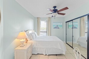 A bedroom with a bed, a nightstand with a lamp, a ceiling fan, and a painting of a pier. - Beachfront Condo at Windward Pointe! (Orange Beach)