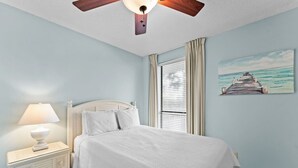 A bedroom with a bed, a nightstand, a ceiling fan, a window with curtains, and a painting of a pier.