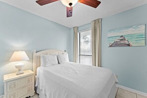 A bedroom with a bed, a nightstand, a ceiling fan, a window with curtains, and a painting of a pier. - Beachfront Condo at Windward Pointe! (Orange Beach)