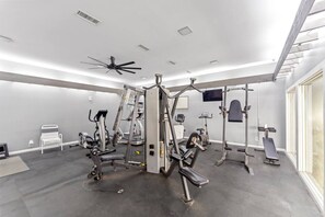 Academia com ventiladores de teto, diversos aparelhos de ginástica e uma grande janela.