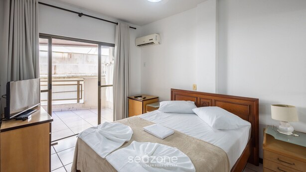 A hotel room with a bed, a television, a desk, a nightstand, and a ceiling fan. - Apt w/ Balcony - Seafront in Meia Praia Ceo0104 (Itapema)