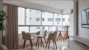 A modern living room with a large window, a sofa, a dining table with chairs, and a TV mounted on the wall.
