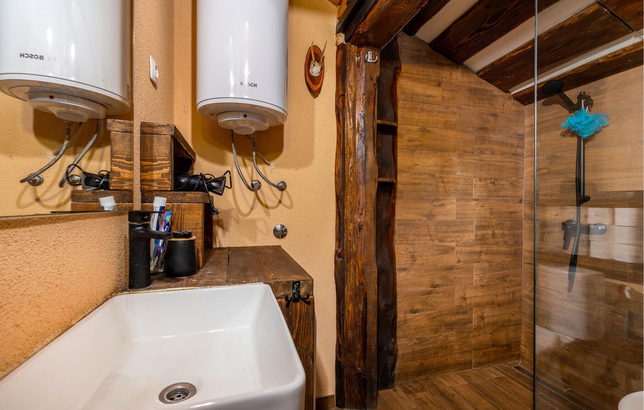 Ein Badezimmer mit einem weißen Waschbecken, zwei an der Wand montierten Bosch-Haartrocknern, einem Holzregal mit Toilettenartikeln und einer Duschkabine aus Glas.