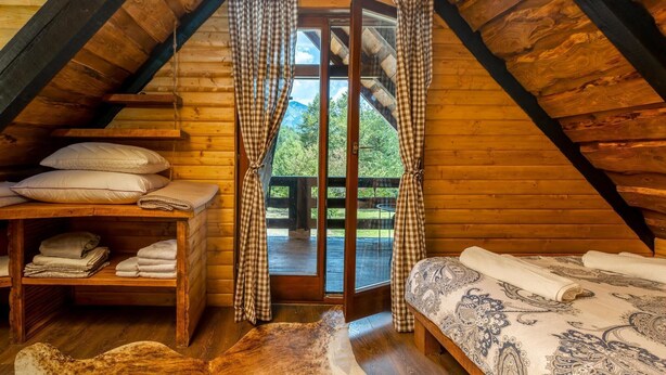 A cozy attic bedroom with a wooden sloped ceiling, a bed with patterned bedding, a shelf with folded towels, and a view of greenery outside.