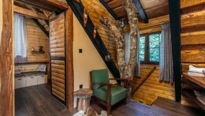 A rustic cabin interior with wooden walls, a green chair, a wooden ladder, and a window with a view of trees.