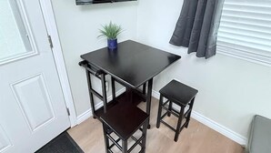 Una habitación compacta con una mesa de madera oscura y dos sillas a juego, una pequeña planta en maceta, un televisor de pantalla plana montado en la pared y una cortina gris.