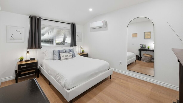 Un dormitorio moderno con una cama grande, mesitas de noche, un equipo de aire acondicionado, un espejo y una ventana con persianas.