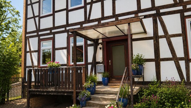 Un bâtiment à colombages avec un escalier en bois menant à une porte, le tout entouré de plantes en pot et d’un chemin en briques.