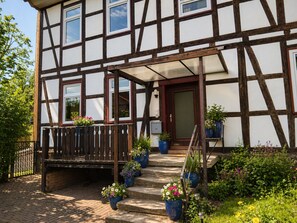 Ein Fachwerkhäuschen mit einer Holztreppe, die zu einer Tür führt, umgeben von Kübelpflanzen und einem Ziegelweg.