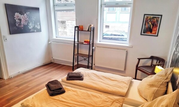 Ein Zimmer mit zwei Betten, einem Heizkörper, einem Fenster mit Blick auf die Straße und einem Bücherregal.