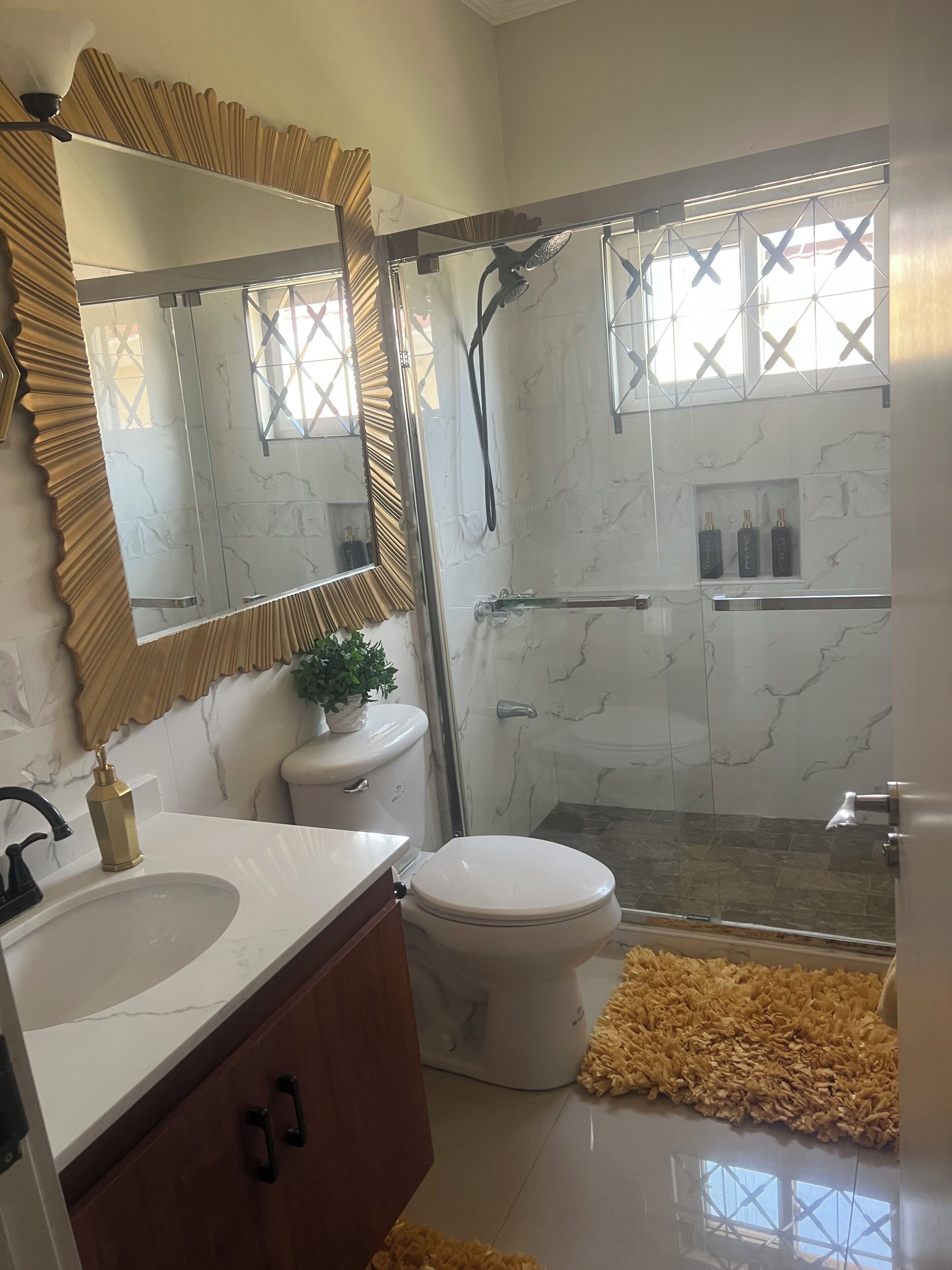 Ein Badezimmer mit einem weißen Marmorwaschbecken, einer Toilette, einer Dusche mit Glastüren und einem großen Spiegel mit Goldrahmen.