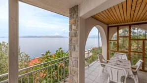 A balcony with a view of a lake and mountains, white outdoor furniture, and stone pillars.