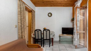 A small, enclosed balcony with a wooden ceiling, a sofa, two chairs, a small table with a TV, and a glass door.