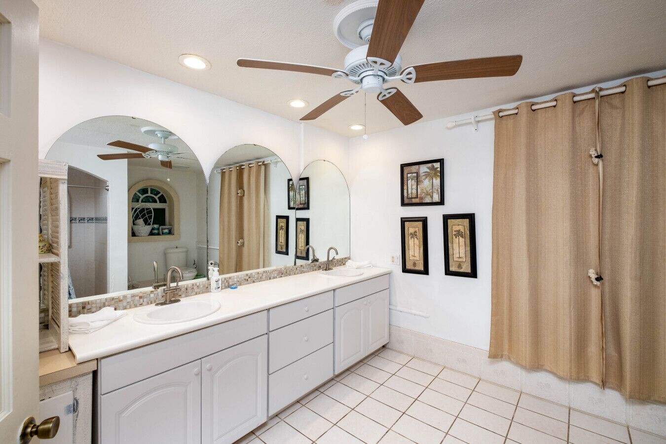 A spacious bathroom with a large vanity, two sinks, a ceiling fan, and a shower area.