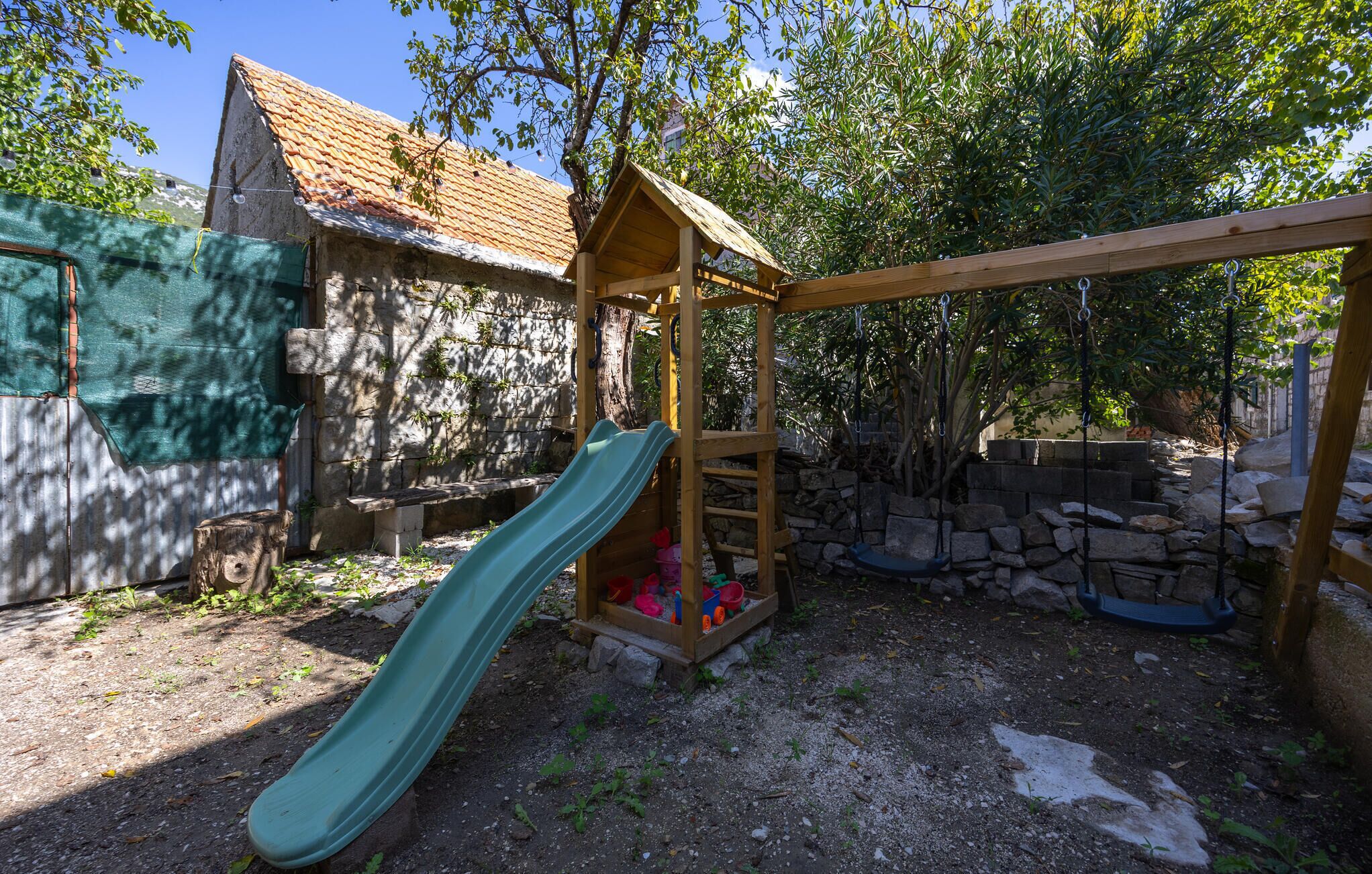 A wooden play structure with a slide, surrounded by a stone wall and trees.