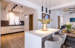 A modern kitchen with a central island, a dining area with grey chairs, and a living space with a sofa and a coffee table.