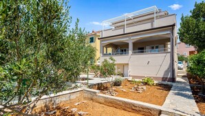 Un bâtiment à deux étages doté d’un balcon, d’un jardin avec des plantes et un petit arbre, ainsi qu’un chemin pavé menant à l’entrée.