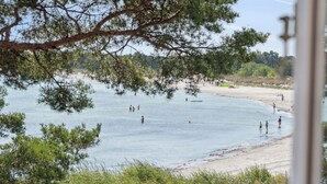 Une plage où l’on voit des gens se baigner et se promener le long du rivage, entourée d’arbres et sous un ciel dégagé.