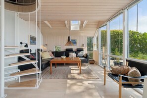 Un salon moderne avec un plafond en bois, un canapĂ© noir, une table basse en bois et de grandes fenĂȘtres donnant sur un paysage verdoyant.