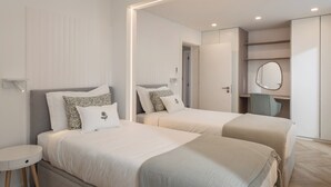 A modern hotel room with two beds, a small desk, a mirror, and built-in shelving.