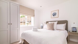 A bedroom with a large bed, a window with a beige blind, a bedside table with a lamp, and a framed picture on the wall.