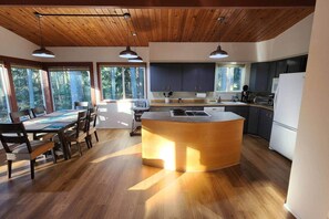 A modern kitchen with a central island, wooden flooring, and large windows offering a view of the outdoors.