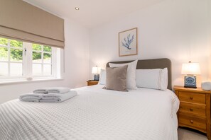 A neatly made bed with white linens and a folded towel, a wooden nightstand with a lamp, and a window with a view of greenery. - Sag Harbour, Sandsend Retreats at Saltmoore - Sag Harbour (Sandsend)