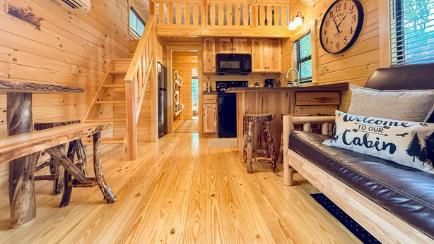 A cabin interior with a wooden staircase, a kitchen area, a seating area with a 'Welcome to our Cabin' pillow, and a clock on the wall. - SaunaHaus | Log Cabin + Private Sauna by the Brook (Lincoln)