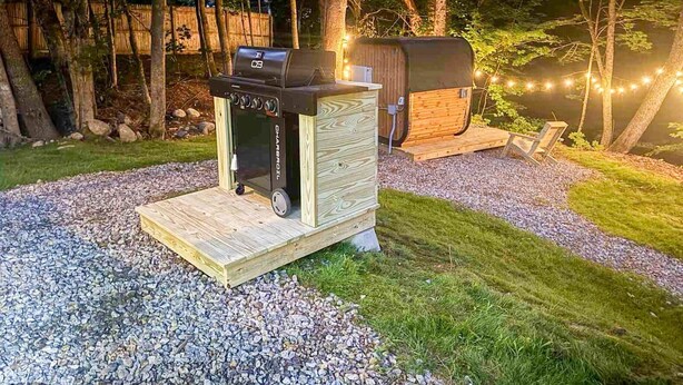Un barbecue portatif doté d’un panneau de commande, d’une terrasse en bois et d’un espace de rangement couvert, le tout situé dans un cadre boisé.