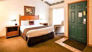 A hotel room with a bed, a nightstand, a chair, a door, and a painting on the wall. - Getaways at Lagonita Lodge (Big Bear Lake)