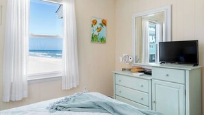 Une chambre à coucher comprenant un lit, une commode, un téléviseur, un tableau représentant des fleurs et une fenêtre donnant sur la plage.