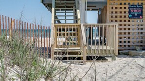 Un bâtiment situé en bord de mer, avec un escalier en bois menant à un balcon, une enseigne indiquant « Location de vacances – L’île vous attend », et une plage en arrière-plan.
