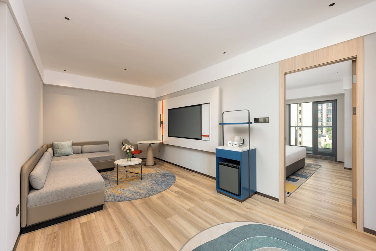 A modern hotel room with a sofa, a small table, a TV, a mini-fridge, and a large mirror.