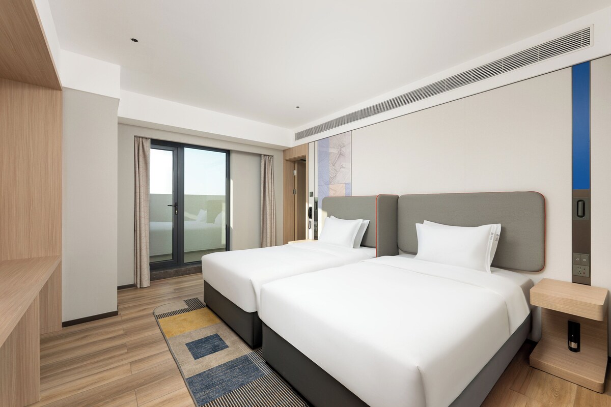 A modern hotel room with two beds, a wooden floor, a large window, and a sliding door leading to a balcony.