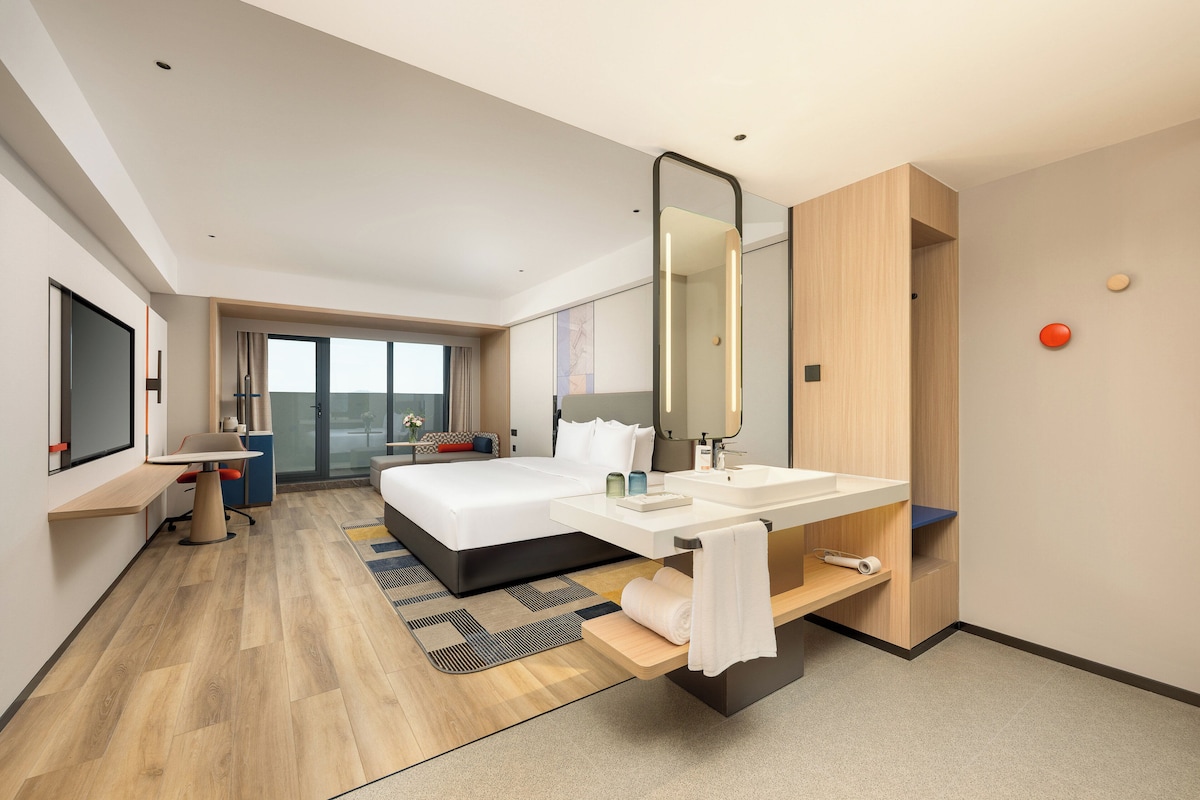 A modern hotel room with a large bed, a desk, a desk chair, a large mirror, a vanity with a sink, and a view of the outdoors.