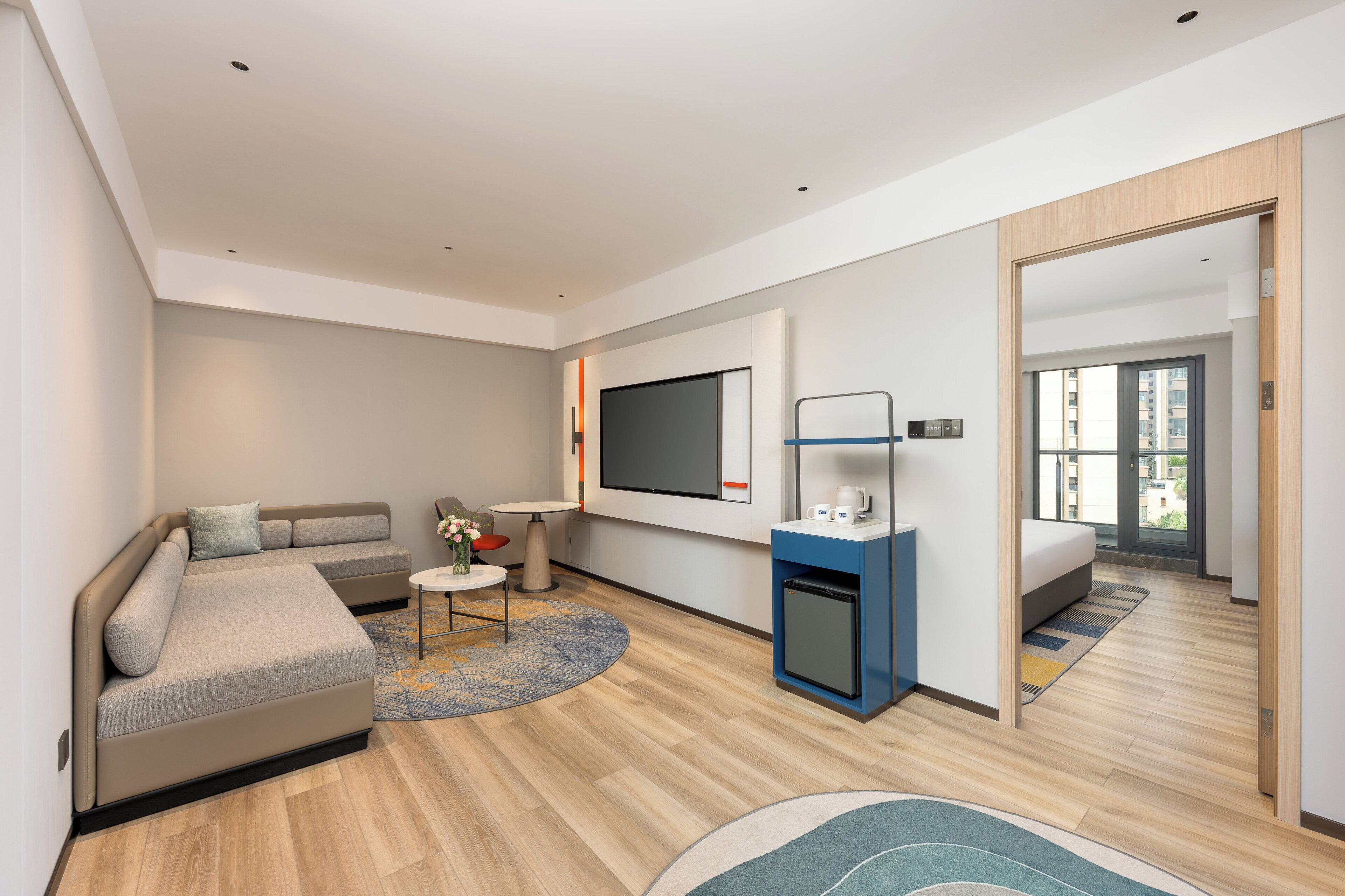 Una habitación de hotel moderna con sofá, una mesita, televisor, refrigerador pequeño y un espejo grande.