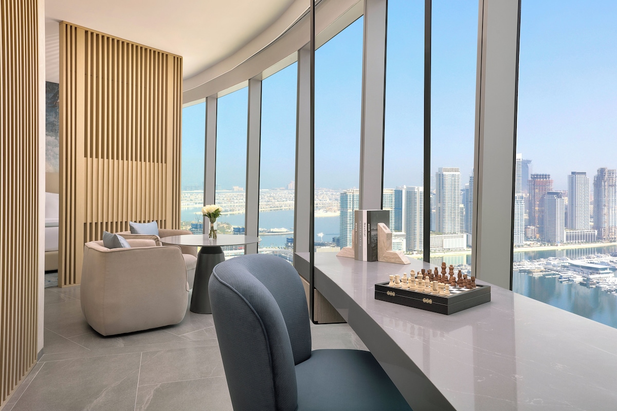 A modern living room with a view of a city skyline and water. It includes a sofa, a chair, a table with a chessboard, and a decorative wooden panel.