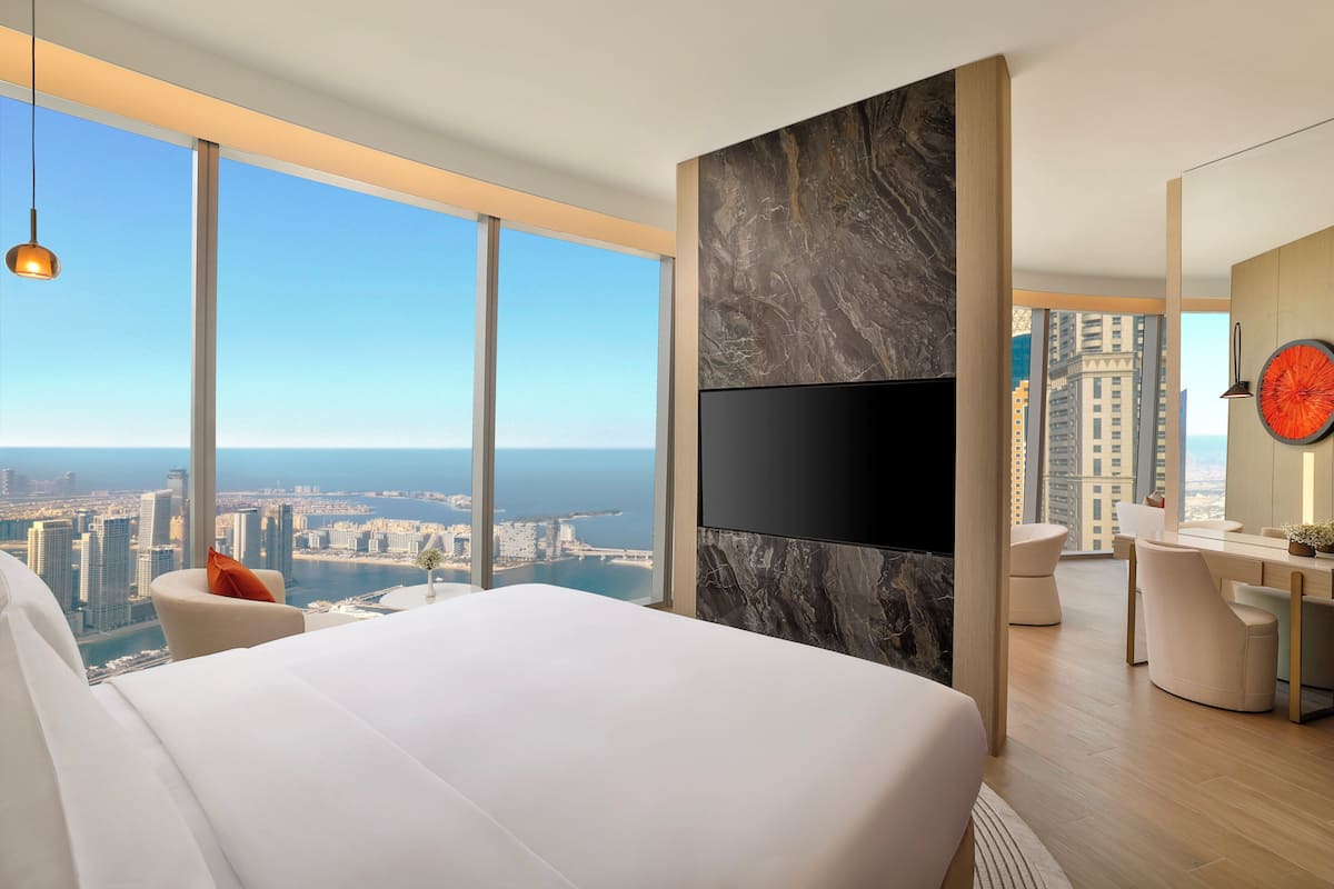 A modern hotel room with a large bed, a desk, and a TV. The room has a city view through large windows.