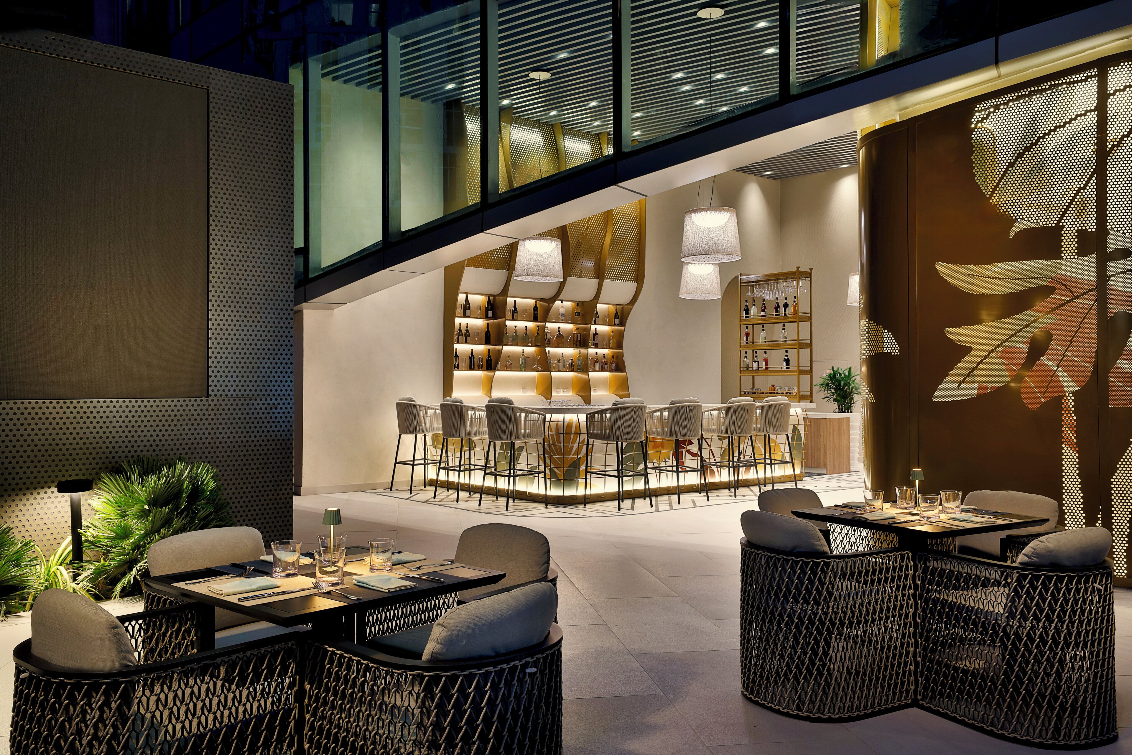 An outdoor seating area with wicker furniture and a bar/lounge area with shelves of bottles and a large mural.