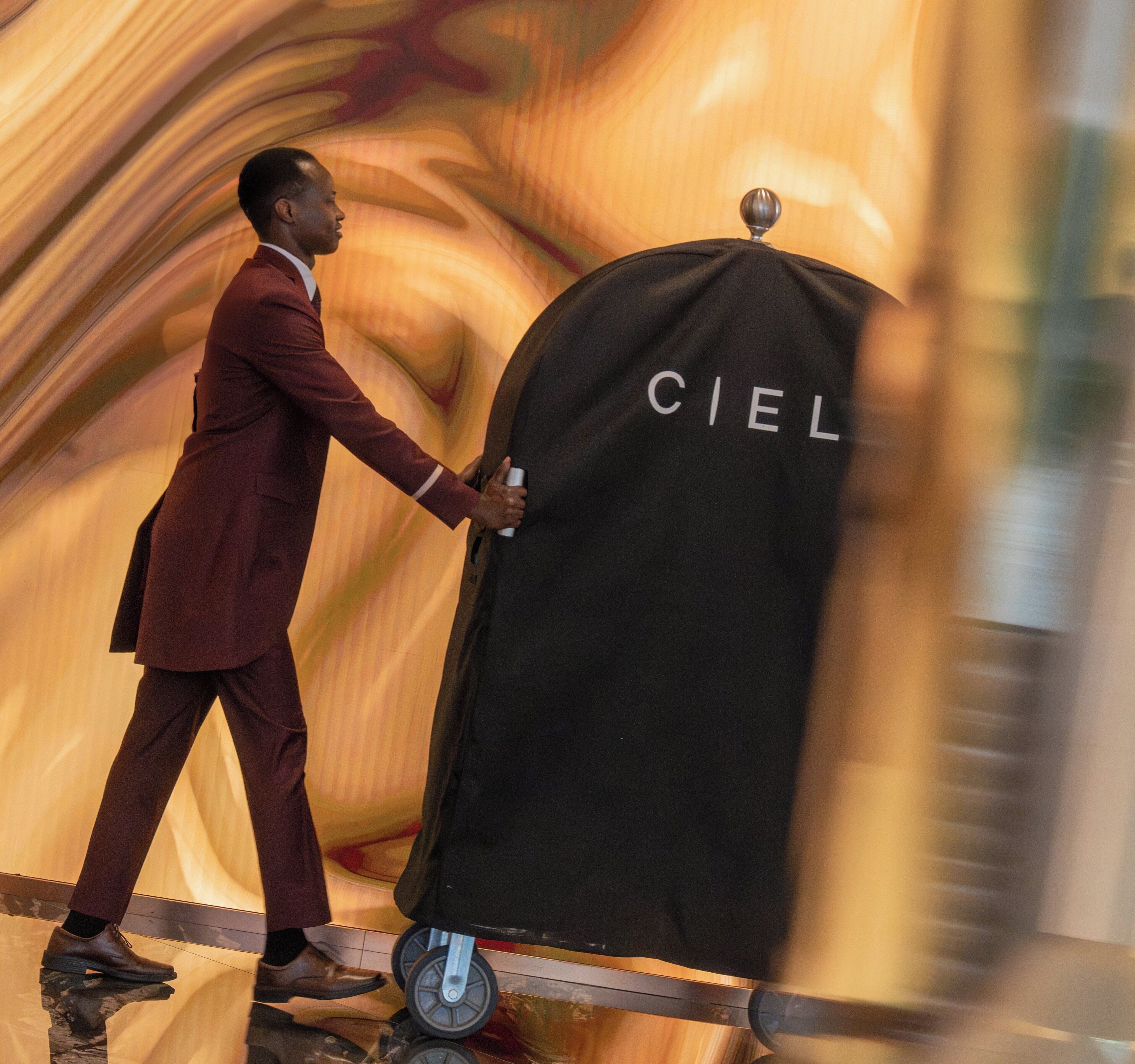 A person pushing a luggage cart with a black cover labeled 'CIEL' in a corridor with a textured wall.