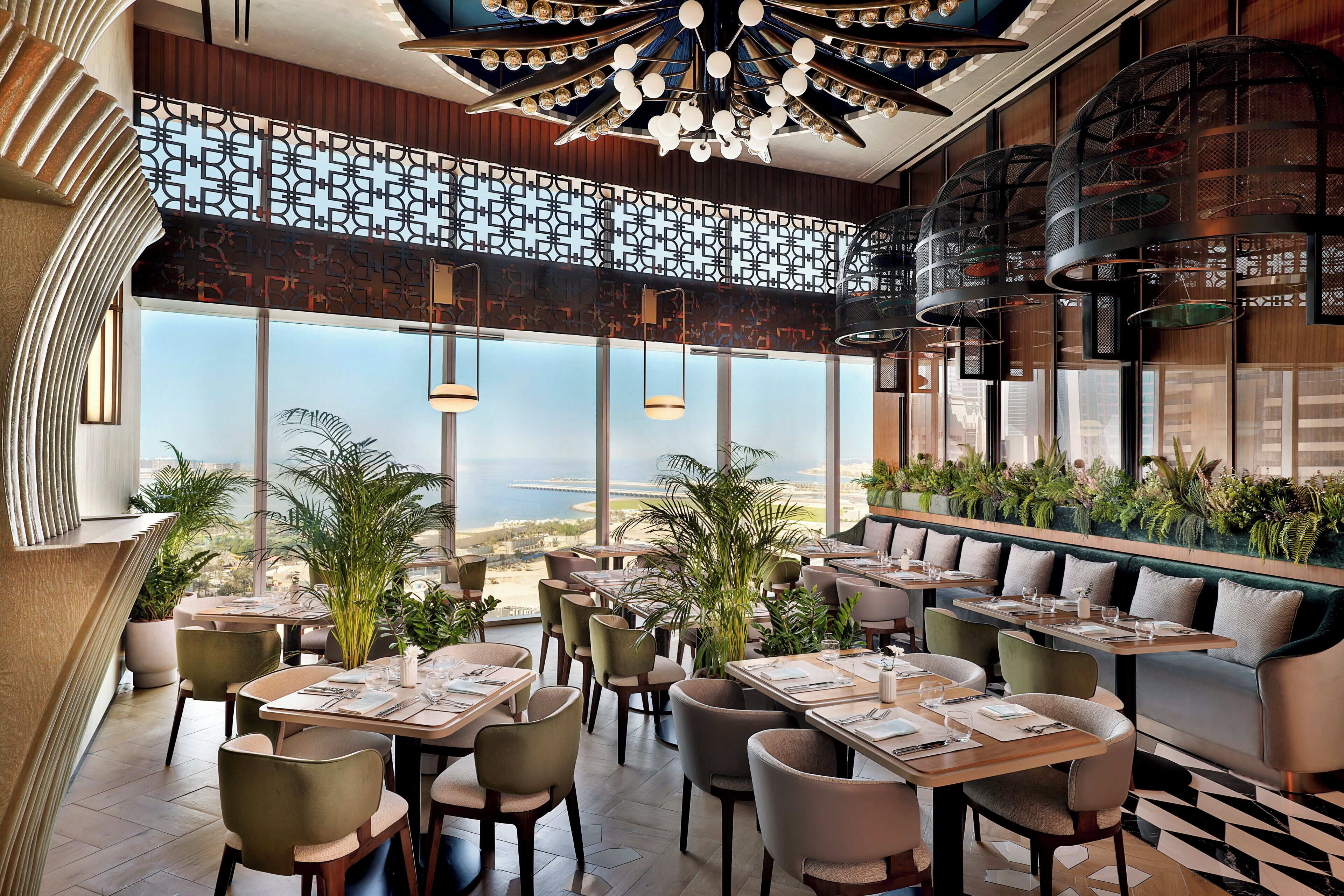 A restaurant with a view of the sea, featuring tables and chairs, a high ceiling with a chandelier, and a patterned wall.