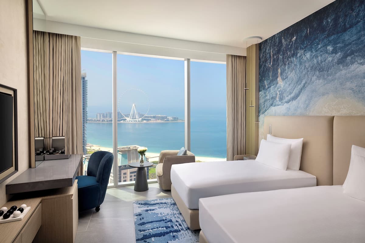 A hotel room with two beds, a TV, a desk, and a view of the sea and a Ferris wheel.