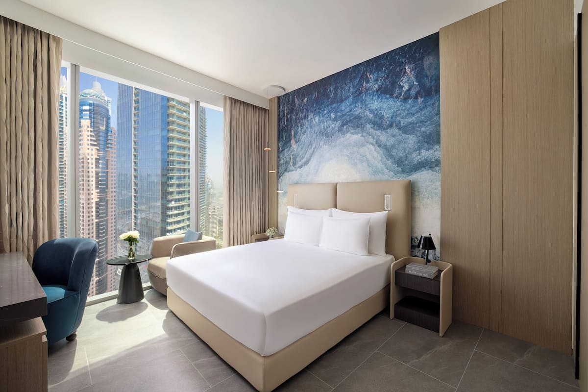 A modern hotel room with a large bed, a desk, a chair, a small table with a vase, and a nightstand with a lamp.