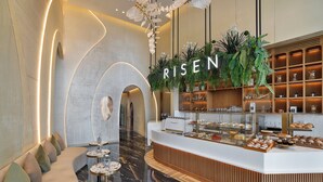 Una zona de recepción moderna con un espacio de asientos curvo, un mostrador con pasteles y un letrero 'RISE' sobre una exhibición de plantas.