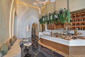 A modern reception area with a curved seating area, a counter displaying pastries, and a 'RISE' sign above a plant display. - Ciel Dubai Marina, Vignette Collection by IHG (Dubai)