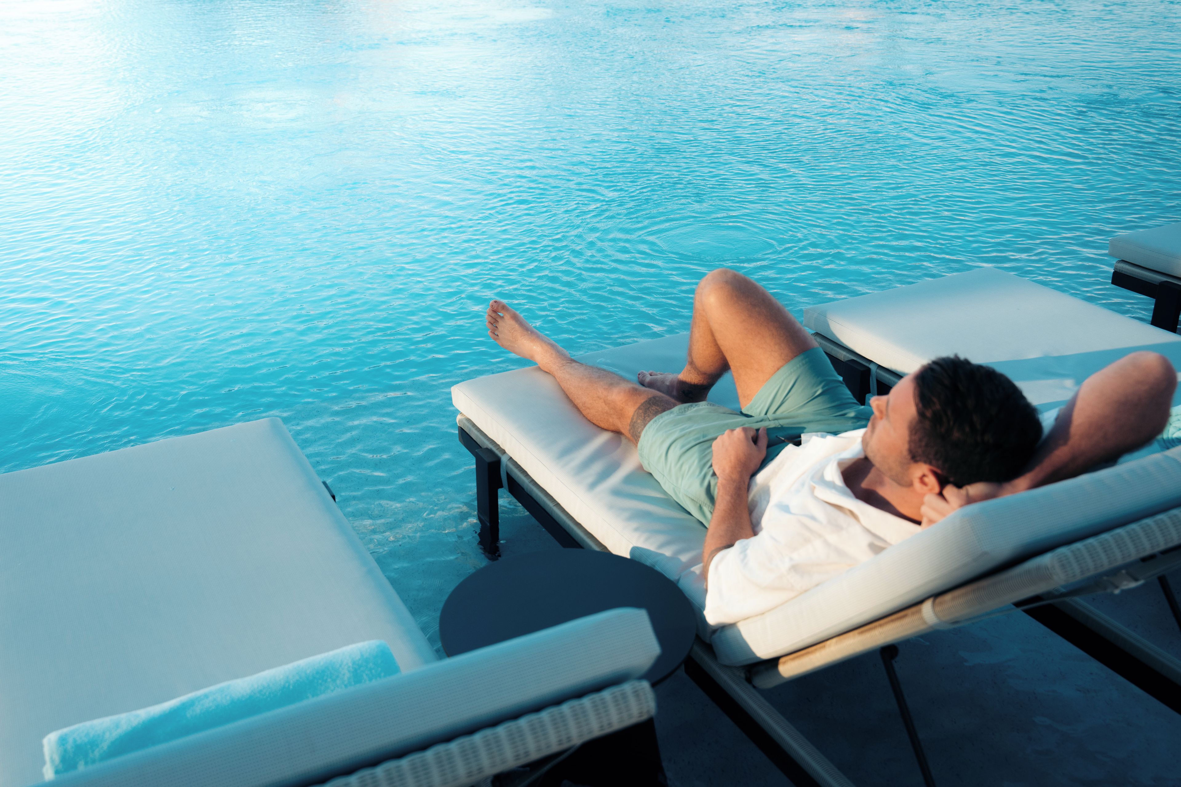 A person reclining on a sun lounger by a pool with clear blue water.