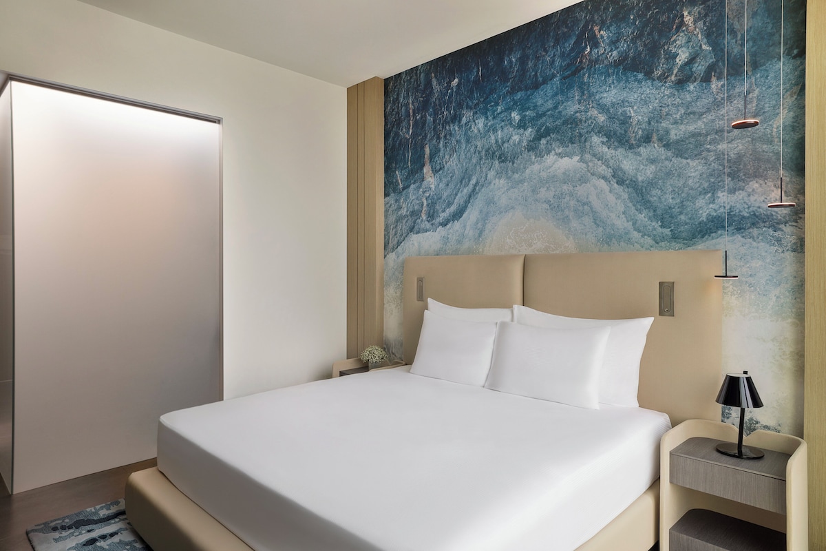 Ein modernes Hotelzimmer mit einem großen Bett, einem Nachttisch mit Lampe und Wandbild.