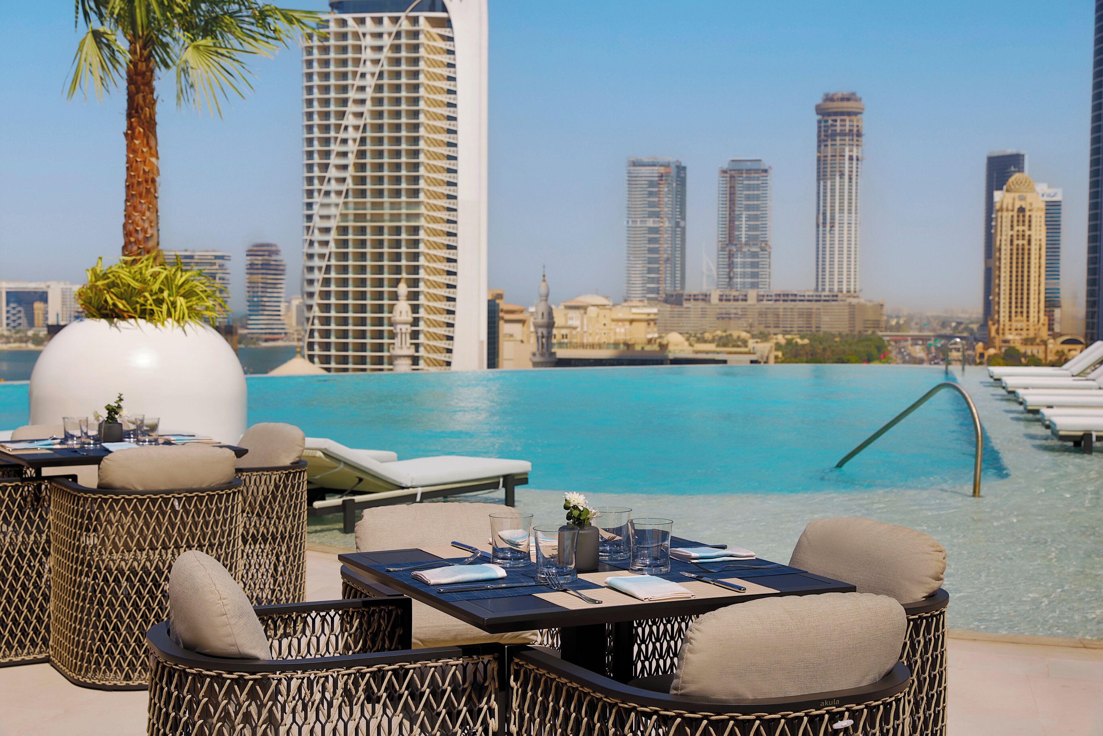 A poolside area with wicker furniture, a table set for dining, and a cityscape view in the background.